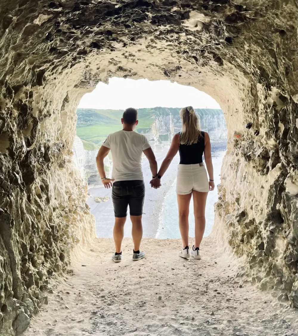 Julian und Ricci stehen Hand in Hand vor einem Felsbogen am Meer